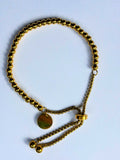 A gold-toned beaded bracelet with a round disc charm and adjustable chain ties, displayed on a light blue background.